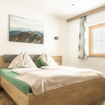 Doppelzimmer mit Holzbett, Nachttisch und Fenster mit Vorhängen.