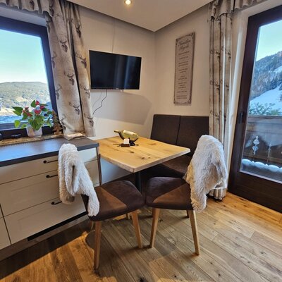 Essbereich der Ferienwohnung mit Eckbank, Holztisch, Fernseher und Ausblick auf den See sowie die schneebedeckte Skipiste vom Balkon.