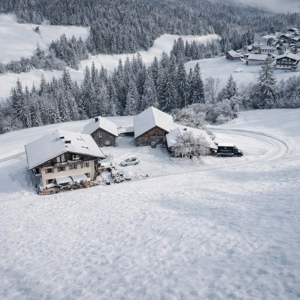 Der Bauernhof in einer verschneiten Berglandschaft mit umliegenden Gebäuden und einer Zufahrtsstraße.