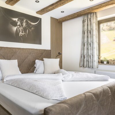 Schlafzimmer der Ferienwohnung mit Doppelbett, großem Bullenbild, Holzdeckenbalken und Fenster mit Blick auf die grüne Landschaft.