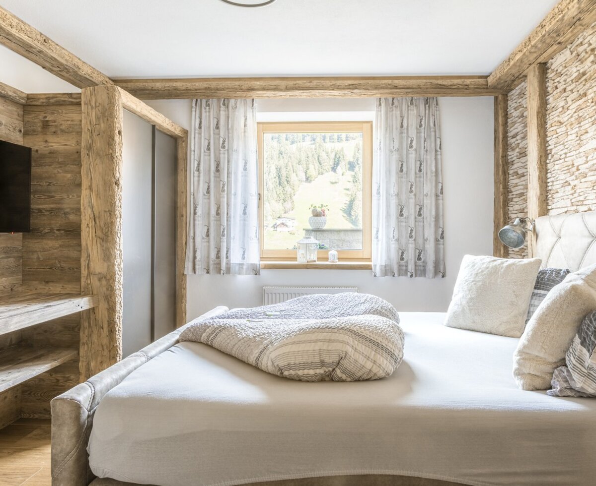Schlafzimmer der Ferienwohnung mit Doppelbett, Holzbalken, Steinwand, integriertem TV und Fenster mit Bergblick.