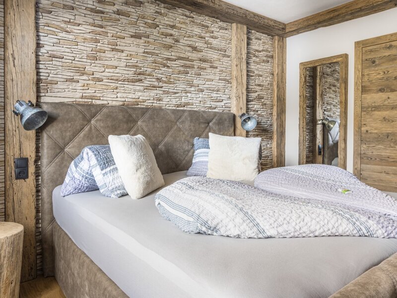 Schlafzimmer der Ferienwohnung mit Doppelbett, gepolstertem Kopfteil, Steinwand, Holzbalken und einem Spiegel.