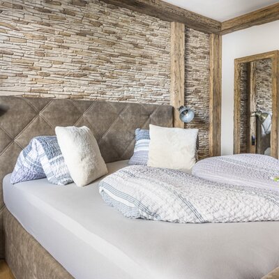 Schlafzimmer der Ferienwohnung mit Doppelbett, gepolstertem Kopfteil, Steinwand, Holzbalken und einem Spiegel.