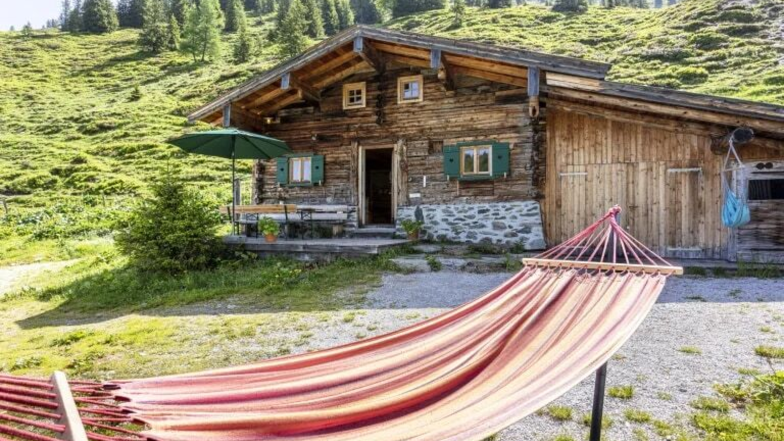 Die Hütte mit einer Holzfassade, einer Terrasse mit Sitzgelegenheiten und Sonnenschirm sowie einer Hängematte im Vordergrund.