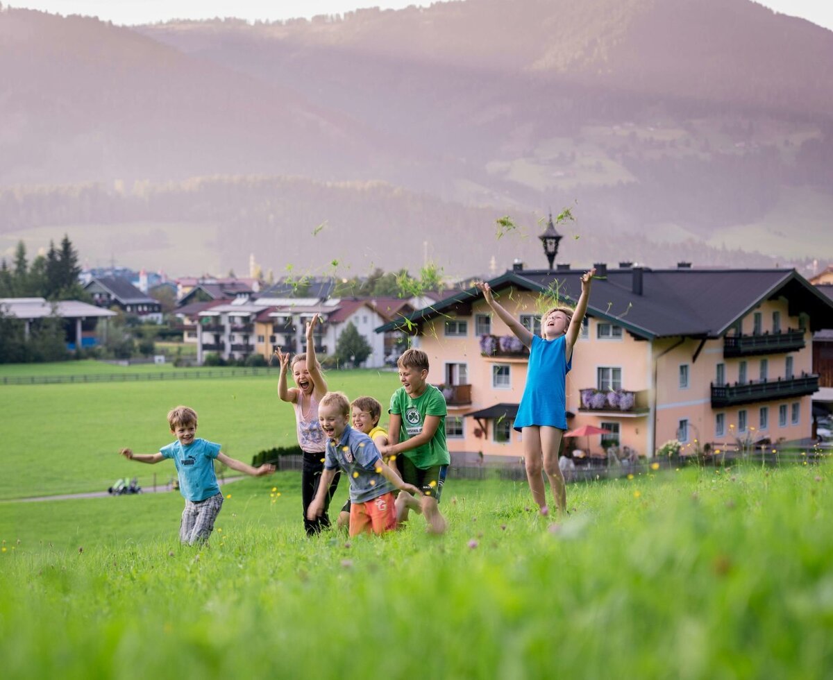 Kinder spielen auf der weitläufigen Wiese des Bauernhofs und werfen Gras in die Luft, mit Blick auf die Berge.