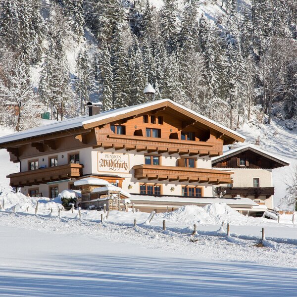 Außenansicht des Bauernhofs "Pension Wildschütz" mit traditioneller Architektur, Balkonen und verschneiter Winterlandschaft.