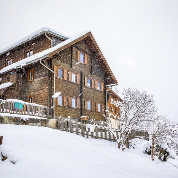 Außenansicht des Bauernhofs im Winter, mit holzverkleideter Fassade und schneebedecktem Dach während des Schneefalls.