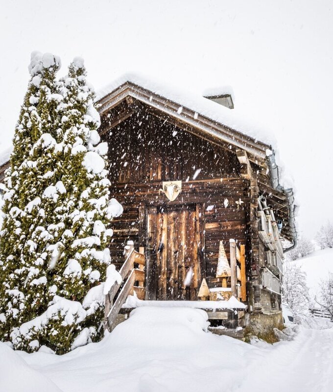 Der Bauernhof in verschneiter Winterlandschaft mit Holzfassade und schneebedecktem Nadelbaum.