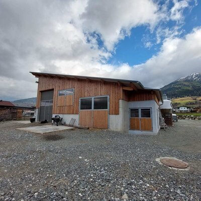 Das landwirtschaftliche Gebäude mit Holzfassade und großen Toren, umgeben von einer Schotterfläche.