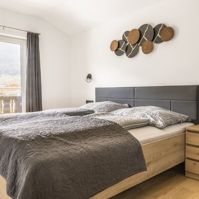 Schlafzimmer der Ferienwohnung mit Doppelbett, Holzbettrahmen, Nachttisch und Zugang zum Balkon mit Bergblick.