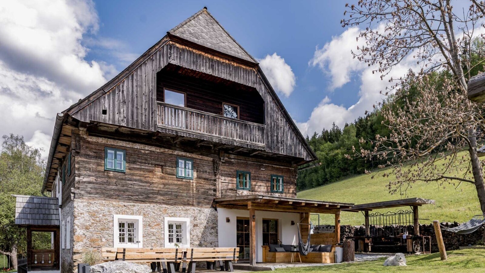 Die Außenseite des Bauernhofs mit traditioneller Holz- und Steinfassade, einem Balkon und einer überdachten Terrasse mit Sitzgelegenheiten im Garten.