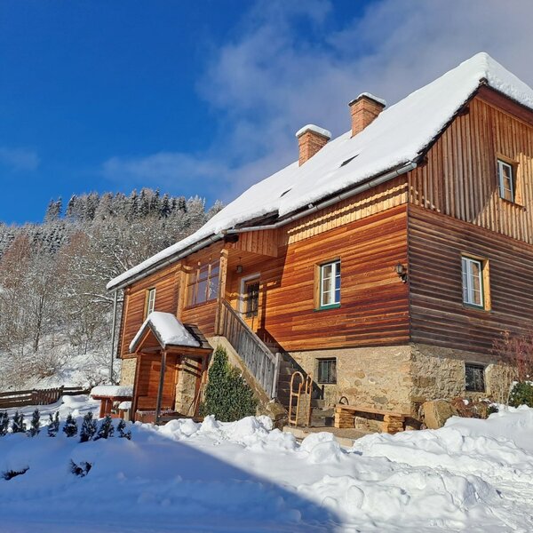 Das Hofgebäude des Almhaus Freitaghube im Winter, mit einer Holzfassade, einem Steinsockel und einem gemauerten Holzkohlegrill im verschneiten Garten.