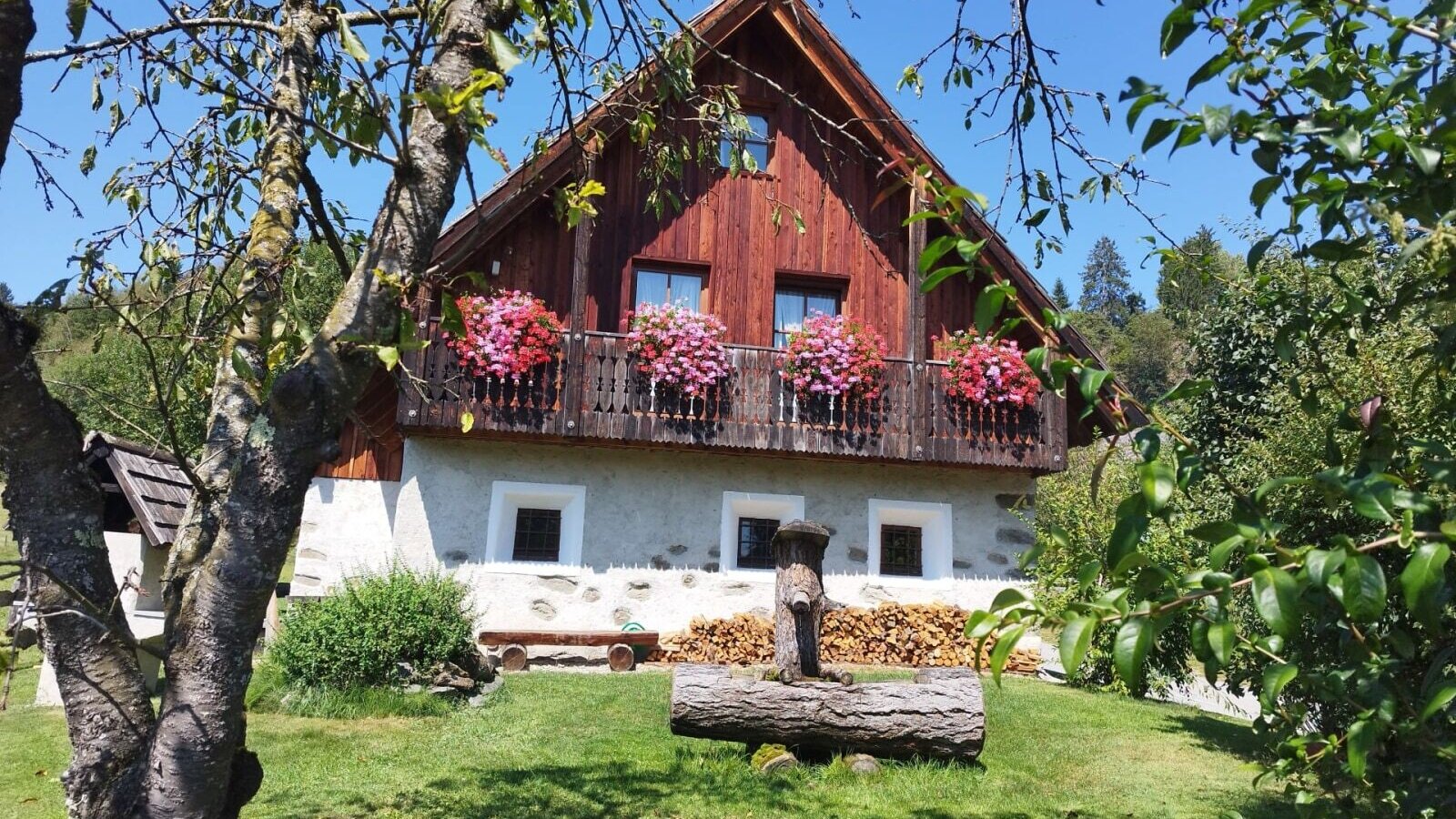 Der einladende Bauernhof mit blumengeschmücktem Balkon und einer grünen Liegewiese mit rustikalem Holzbrunnen.