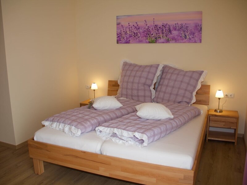 Schlafzimmer mit Doppelbett, lila Bettwäsche, zwei Nachttischen mit Lampen und einem Lavendelbild.