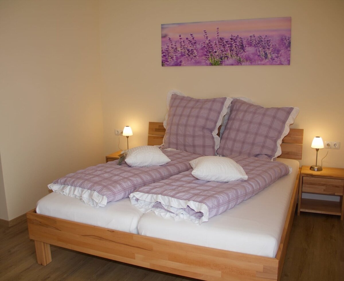 Schlafzimmer mit Doppelbett, lila Bettwäsche, zwei Nachttischen mit Lampen und einem Lavendelbild.