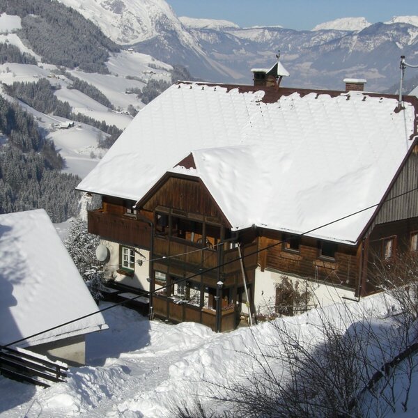 Der Bauernhof mit Holzbalkonen und schneebedecktem Dach, umgeben von einer winterlichen Berglandschaft.