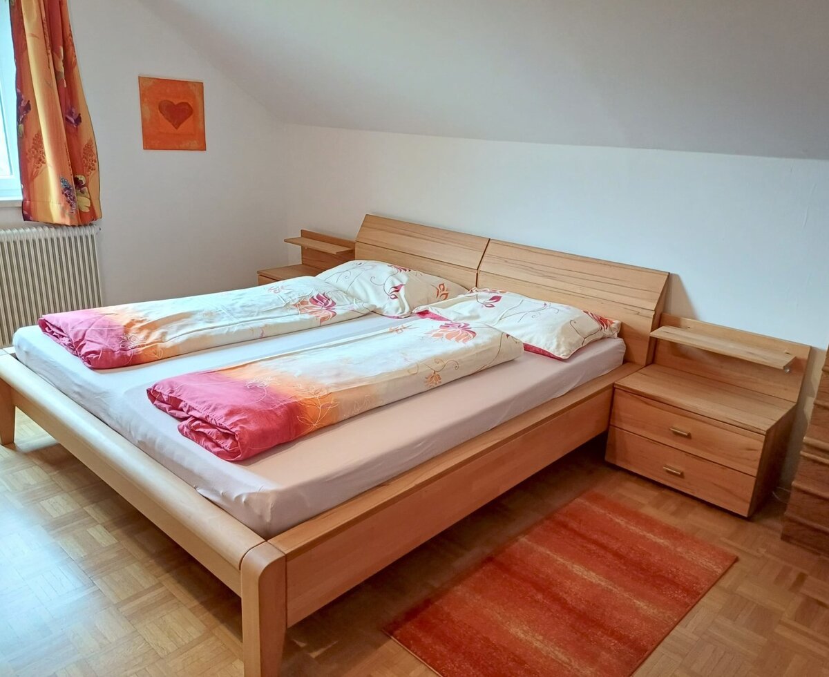 Schlafzimmer im Bauernhof mit Doppelbett, zwei Nachttischen, Fenster mit Vorhängen und Dachschräge.
