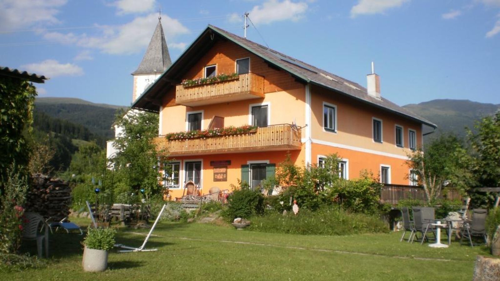 Der Bauernhof mit blumengeschmückten Balkonen, einer Rasenfläche und Blick auf die umliegenden Berge und den Kirchturm.