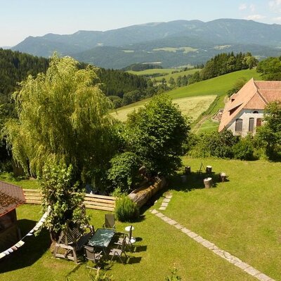 Der Garten des Bauernhofs mit Gartenmöbeln, Hängematte, Pavillon und Blick auf die bewaldeten Berge.