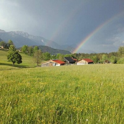 Der Bauernhof umgeben von grünen Wiesen und Bergen, mit einem Regenbogen.