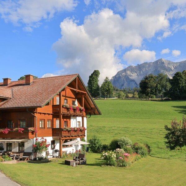 Der einladende Bauernhof mit blumengeschmückten Balkonen inmitten von grünen Wiesen vor einer malerischen Bergkulisse.