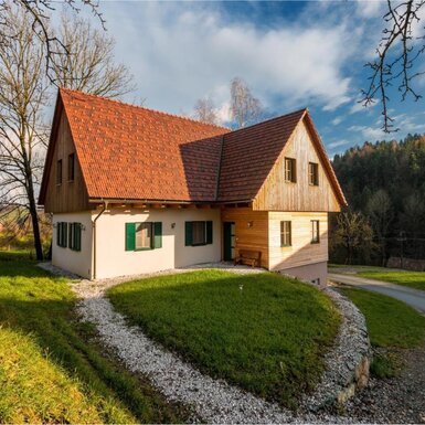 Der Bauernhof von außen, mit rotem Ziegeldach, hellen Wänden, grünen Fensterläden und Holzverkleidungen, umgeben von Rasen und einem Zufahrtsweg.