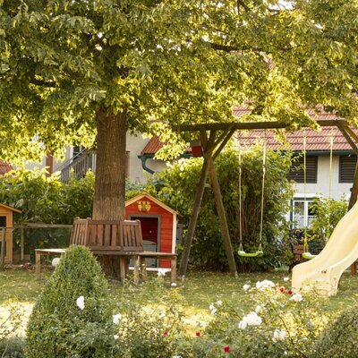 Der einladende Garten des Bauernhofs mit einem schattenspendenden Baum, einer Kinderrutsche, Schaukeln und gemütlichen Sitzecken.