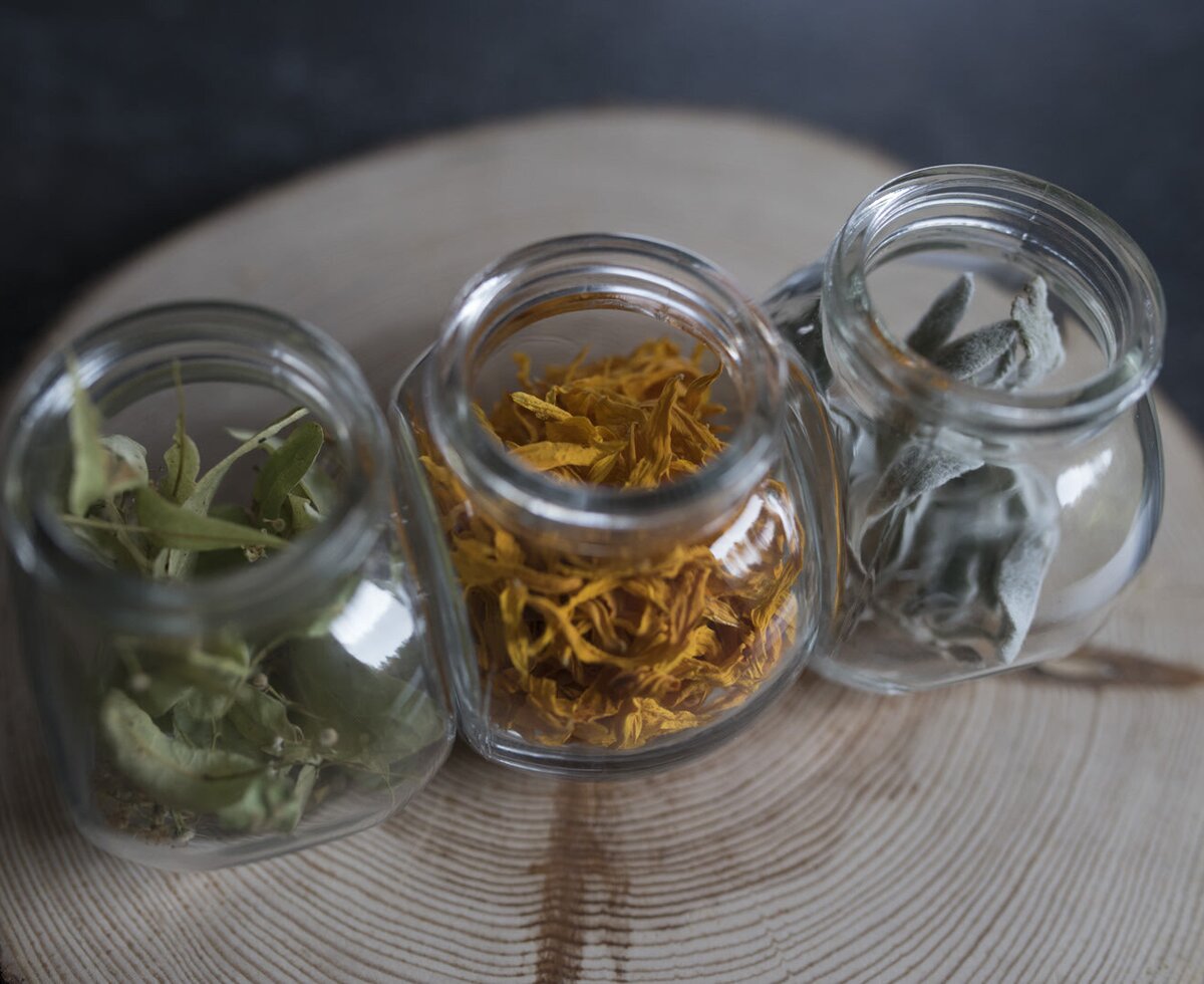 Verschiedene getrocknete Kräuter und Blüten in Gläsern auf einem Holzbrett im Hof.