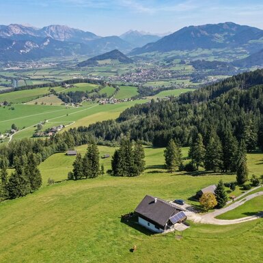 Der Hof liegt auf einem Hügel, umgeben von grünen Wiesen und Wald, mit Panoramablick über das Tal und die Berge.