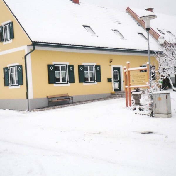Die Außenansicht des Bauernhauses mit gelber Fassade, grünen Fensterläden und einer Sitzbank im Schnee.