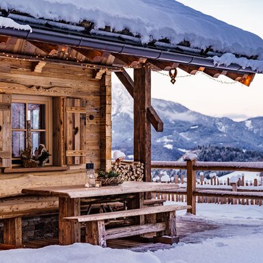 Die schneebedeckte Terrasse des Hofes mit einem Holztisch und Bänken, umgeben von einer winterlichen Berglandschaft.