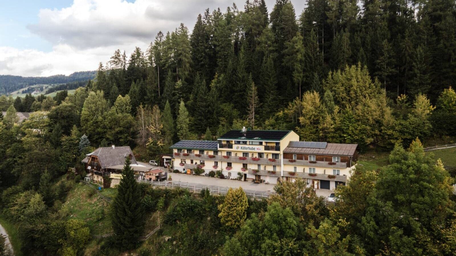 Außenansicht des Bauernhofs Käferhube, bestehend aus mehreren Gebäuden, eingebettet in eine bewaldete Berglandschaft.
