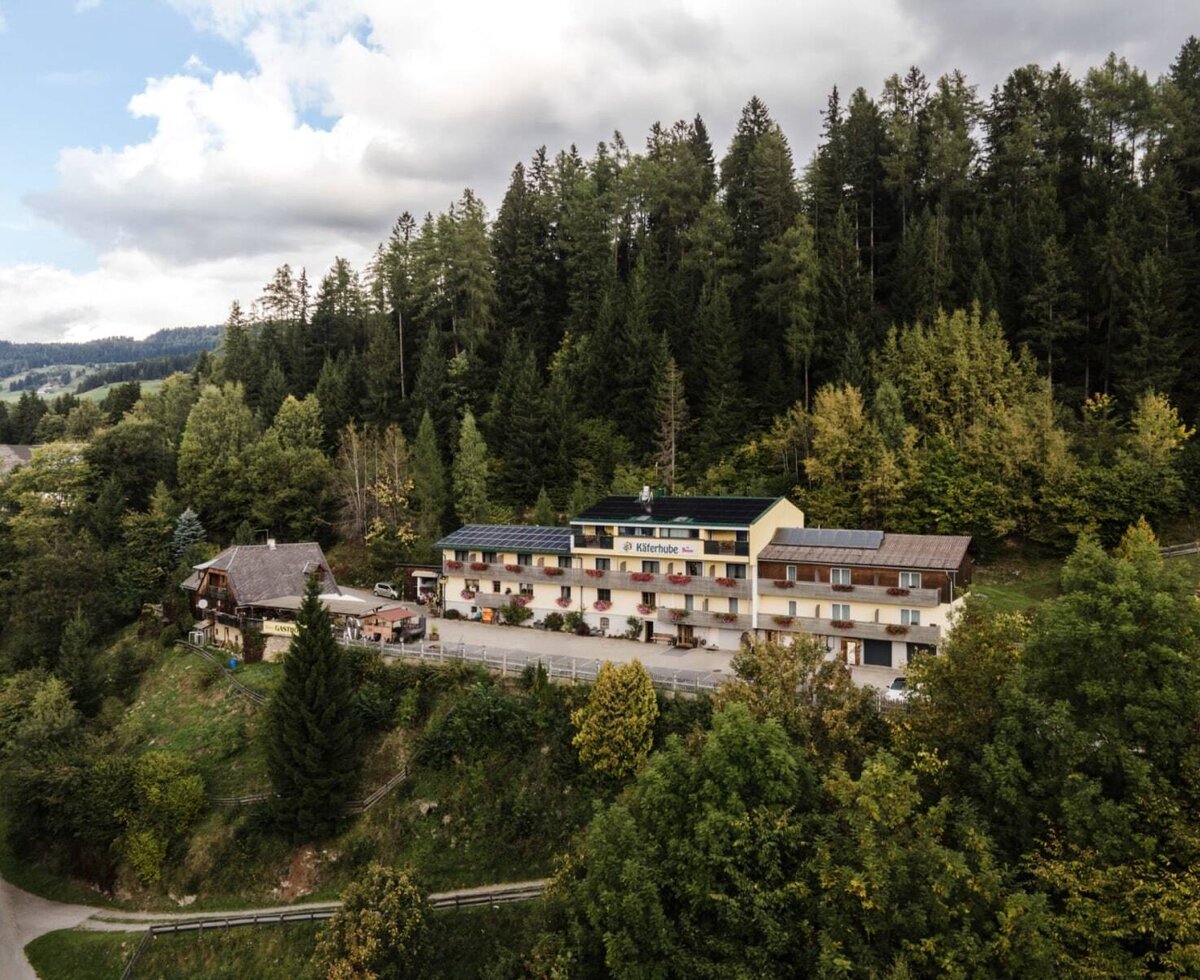 Außenansicht des Bauernhofs Käferhube, bestehend aus mehreren Gebäuden, eingebettet in eine bewaldete Berglandschaft.