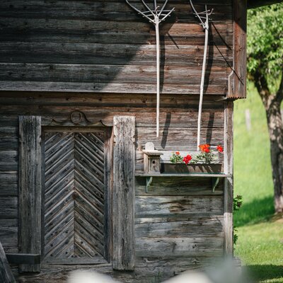 Rustikale Holzwand des Bauernhofs mit zwei Heugabeln, einem Blumenkasten mit roten Blumen und einem Vogelhaus.