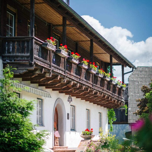 Außenansicht des Bauernhofs mit Holzbalkon, roten Blumen und gewölbtem Eingang.