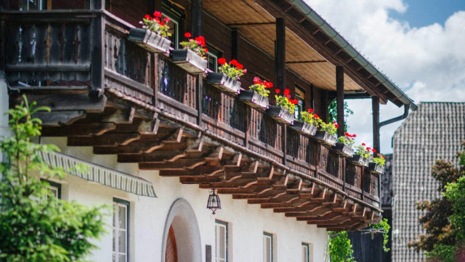 Außenansicht des Bauernhofs mit Holzbalkon, roten Blumen und gewölbtem Eingang.