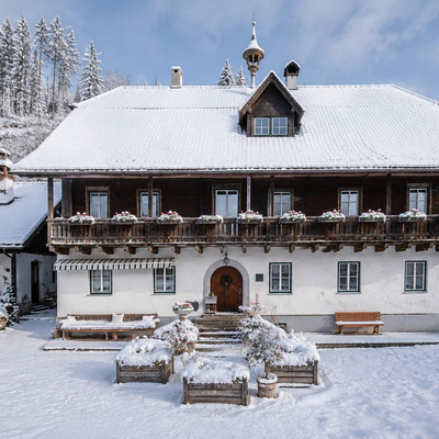 Der traditionelle Bauernhof präsentiert sich im winterlichen Gewand mit schneebedecktem Dach, einem charmanten Holzbalkon und einem verschneiten Vorgarten.