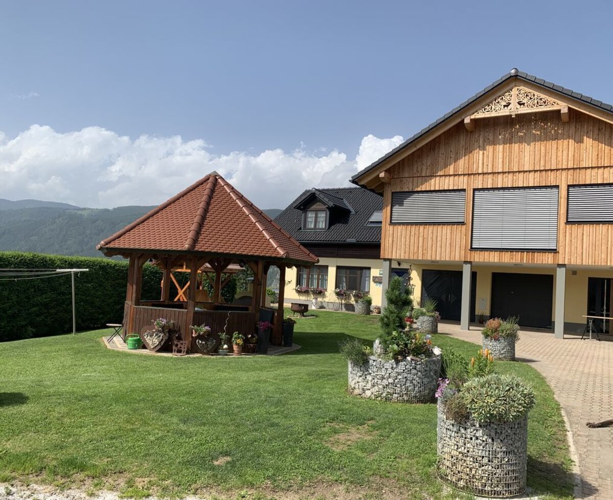Der Bauernhof von außen mit gepflegtem Garten, einem Pavillon und Blick auf die umliegenden Berge.