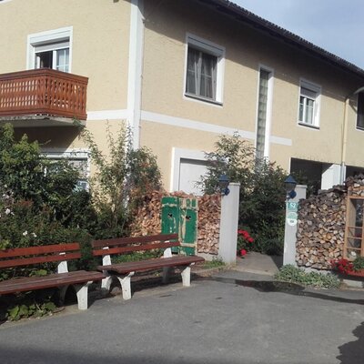 Außenansicht des Bauernhofs mit hellgelber Fassade, Holzbalkon, zwei Sitzbänken und einem Holzstapel am Eingang.