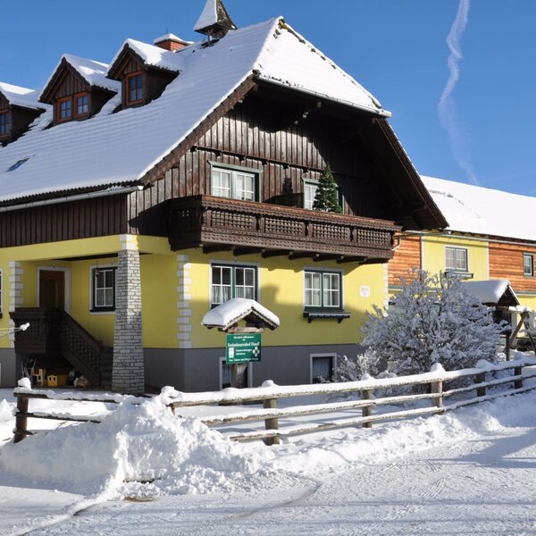 Die Außenansicht des Bauernhofs im Winter, mit gelber Fassade, Holzbalkon und verschneiter Umgebung.