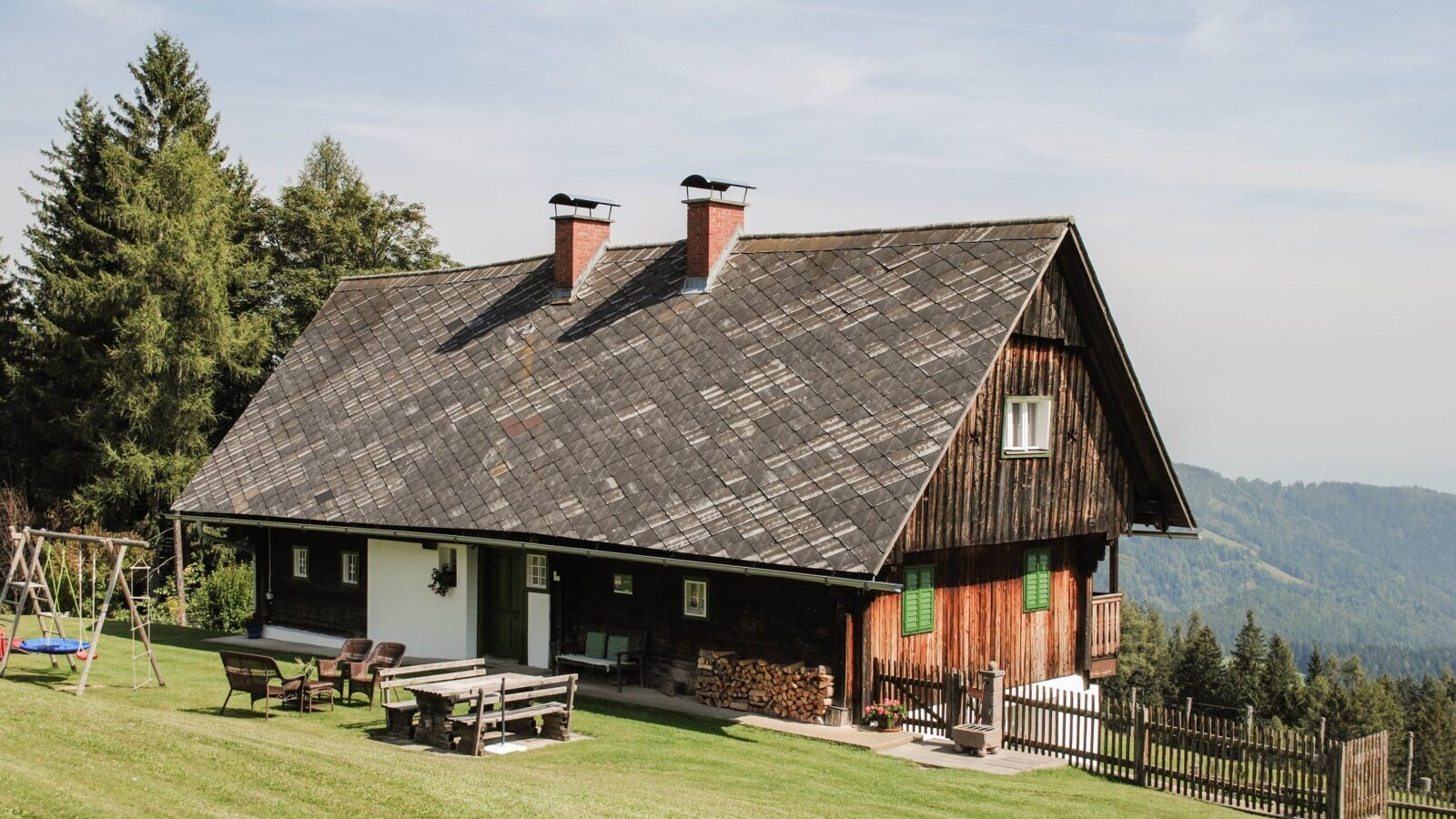Das Bauernhaus, mit Holzfassade, einer Rasenfläche mit Sitzgelegenheiten im Freien, Kinderschaukel und Bergblick.