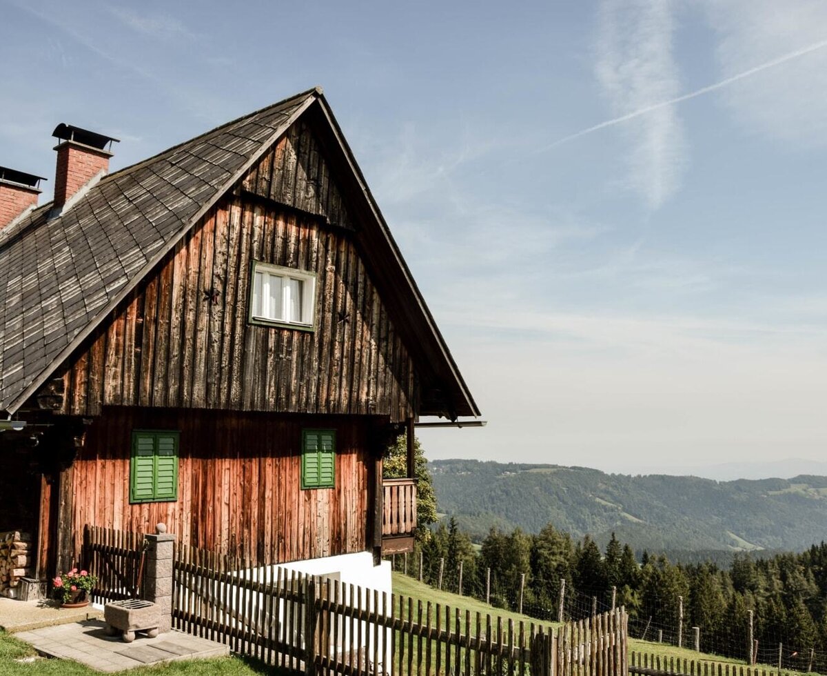 Außenansicht des Bauernhofs mit Holzfassade, traditionellem Dach und Blick auf die bewaldete Berglandschaft.