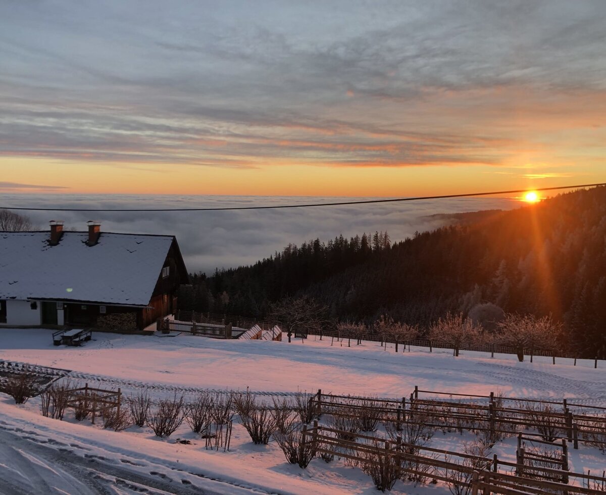 Der schneebedeckte Bauernhof bei Sonnenuntergang über einem Wolkenmeer.