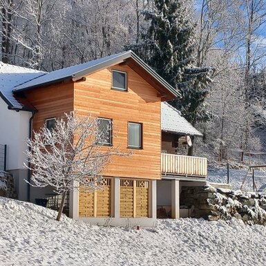 Das Ferienhaus Klampfl im Winter, umgeben von Schnee und Wald, mit einem Balkon und einem Spielbereich.