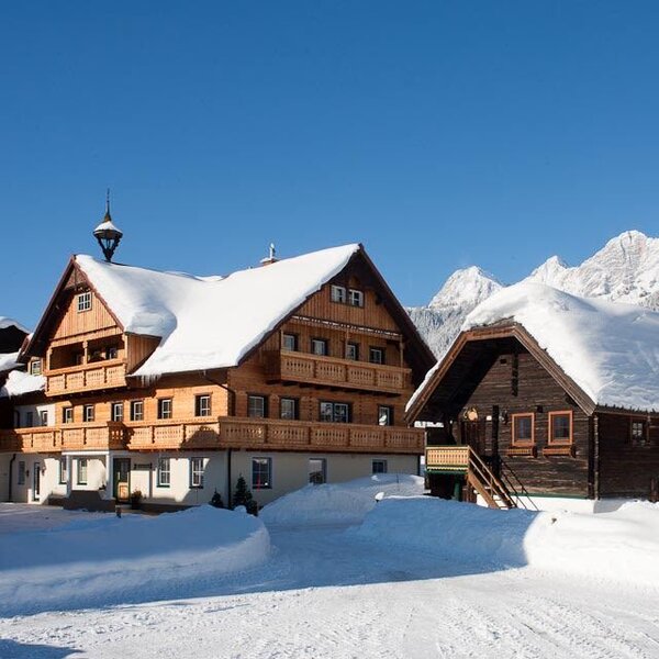 Der Bauernhof im Winter mit schneebedeckten Dächern und Holzbalkonen vor der alpinen Bergkulisse.