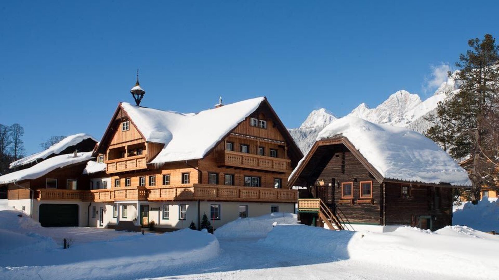Der Bauernhof im Winter mit schneebedeckten Dächern und Holzbalkonen vor der alpinen Bergkulisse.