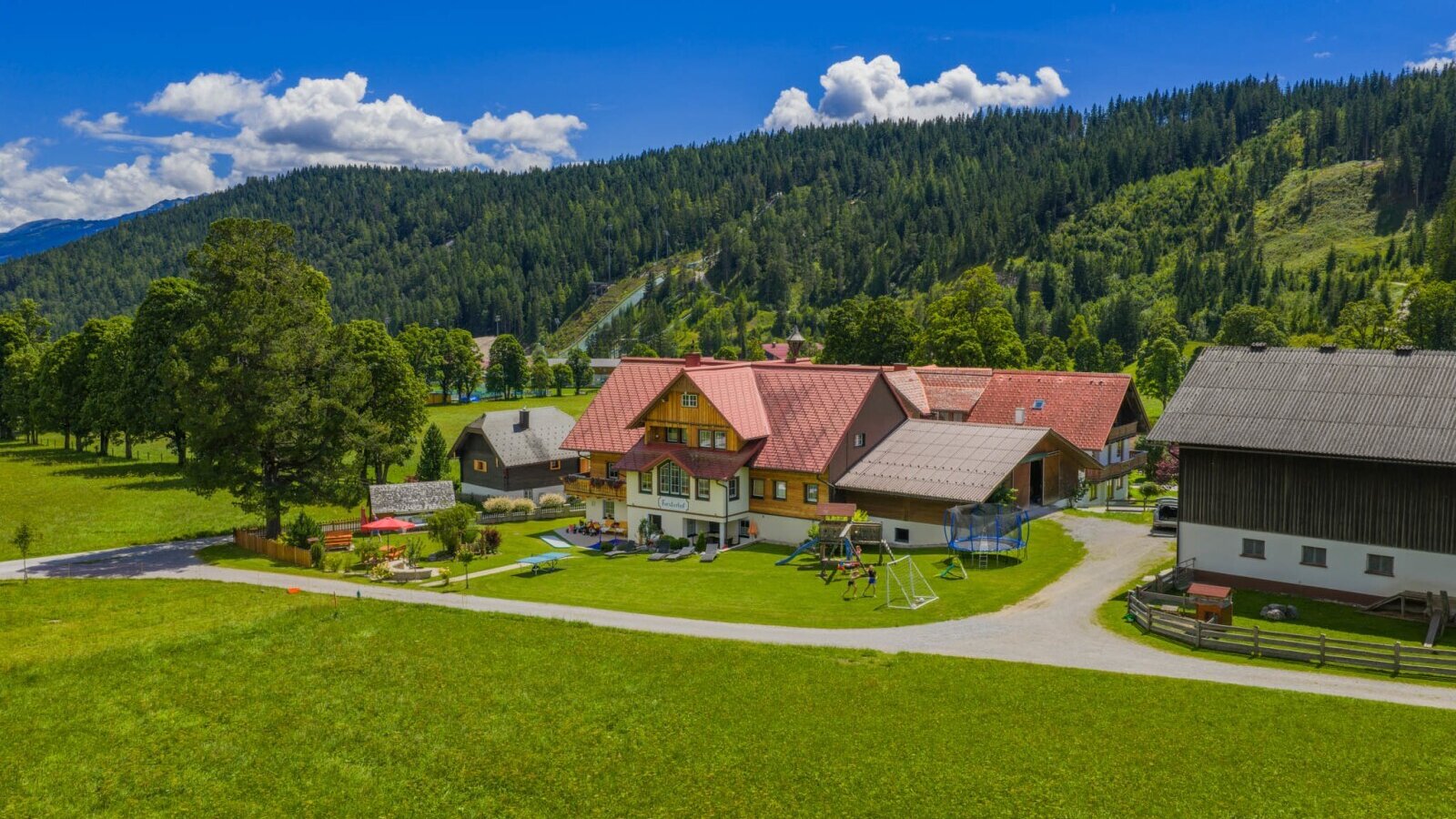 Der Bauernhof mit Spielplatz, Trampolin und Schaukeln vor einer Bergkulisse