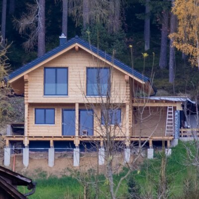Das Bauernhofgebäude ist ein zweistöckiges Holzhaus mit dunklem Dach, gelegen an einem bewaldeten Hang.