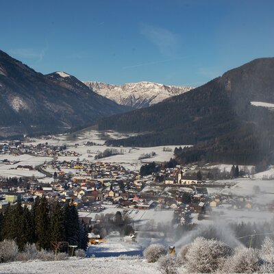 Die verschneite Berglandschaft mit einem Dorf im Tal und einer Skipiste mit aktiven Schneekanonen.