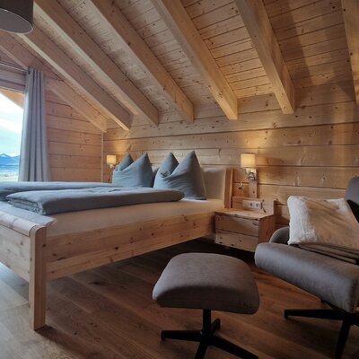 Doppelzimmer im Bauernhof mit Holzvertäfelung, Doppelbett, Sessel, Fußhocker und großer Fensterfront mit Bergblick.